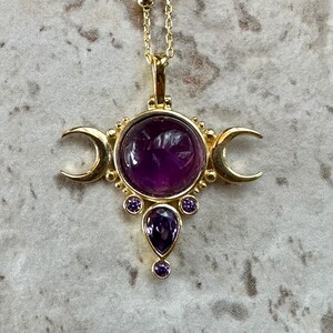 May include: Gold-coloured pendant necklace with a large, round, deep purple gemstone at the centre, flanked by crescent moons. Smaller purple gemstones are set below. The necklace has a delicate chain.