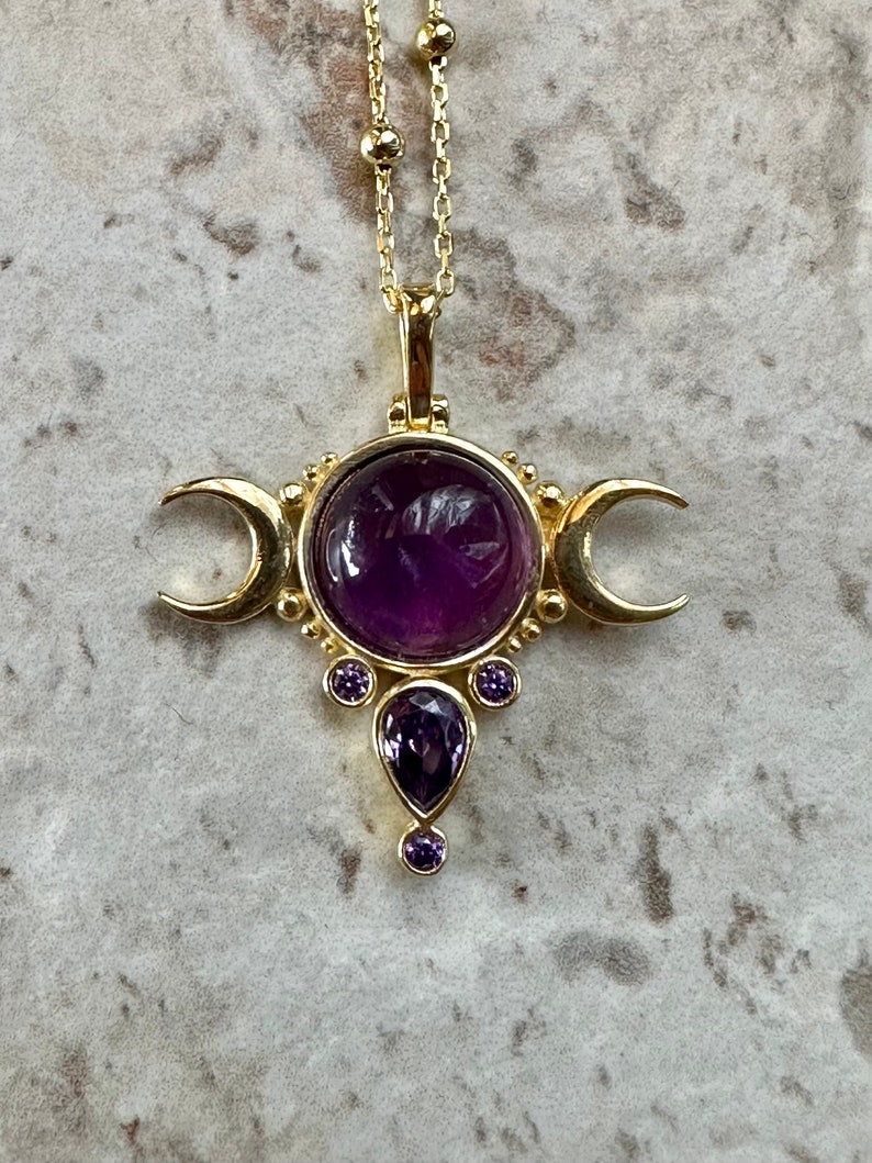 May include: Gold-coloured pendant necklace with a large, round, deep purple gemstone at the centre, flanked by crescent moons. Smaller purple gemstones are set below. The necklace has a delicate chain.