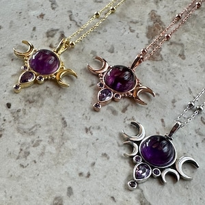 May include: Three necklaces with celestial pendants. Each pendant has a large, round, deep purple gemstone, surrounded by crescent moons and smaller purple stones. The chains are gold, rose gold, and silver.
