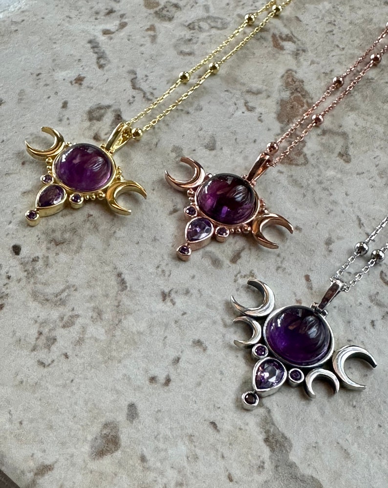 May include: Three necklaces with celestial pendants. Each pendant has a large, round, deep purple gemstone, surrounded by crescent moons and smaller purple stones. The chains are gold, rose gold, and silver.