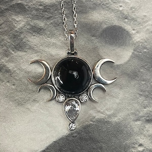 May include: Silver-toned pendant necklace featuring a large, round black stone at the center, surrounded by crescent moon shapes. A teardrop-shaped clear stone and smaller clear stones add sparkle. The necklace hangs on a delicate chain.