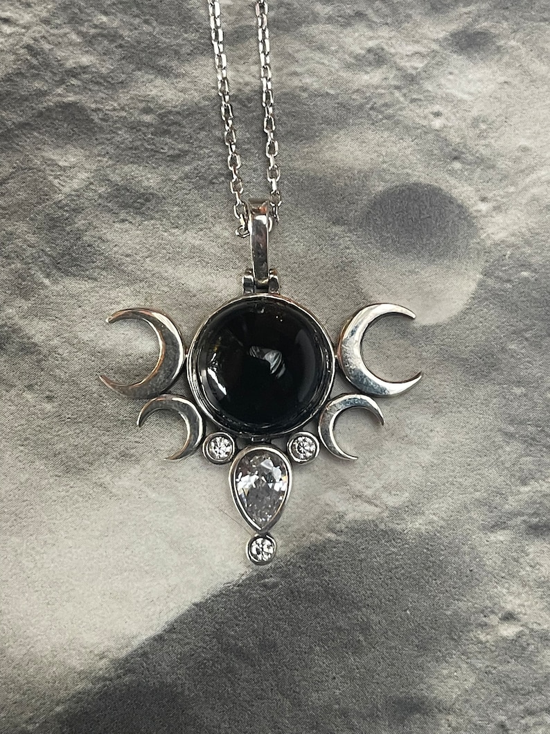 May include: Silver-toned pendant necklace featuring a large, round black stone at the center, surrounded by crescent moon shapes. A teardrop-shaped clear stone and smaller clear stones add sparkle. The necklace hangs on a delicate chain.