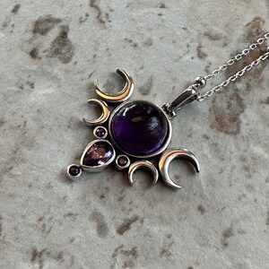 May include: A silver pendant necklace with a large, round, deep purple gemstone at its centre. The pendant is decorated with crescent moon shapes and smaller purple gemstones. The necklace chain is delicate and silver.