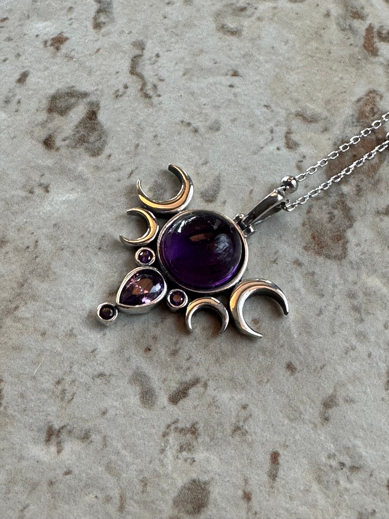 May include: A silver pendant necklace with a large, round, deep purple gemstone at its centre. The pendant is decorated with crescent moon shapes and smaller purple gemstones. The necklace chain is delicate and silver.