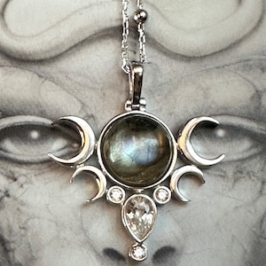 May include: Silver pendant necklace featuring a central iridescent stone, surrounded by crescent moon shapes and clear gemstones. The necklace hangs from a delicate silver chain, showcasing a celestial design.