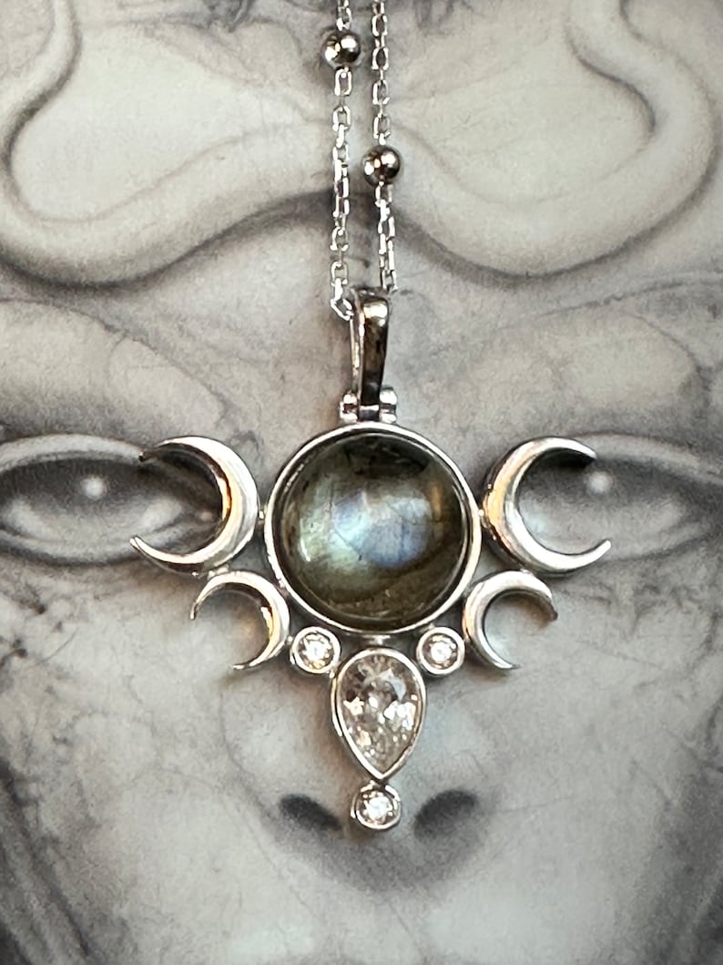 May include: Silver pendant necklace featuring a central iridescent stone, surrounded by crescent moon shapes and clear gemstones. The necklace hangs from a delicate silver chain, showcasing a celestial design.