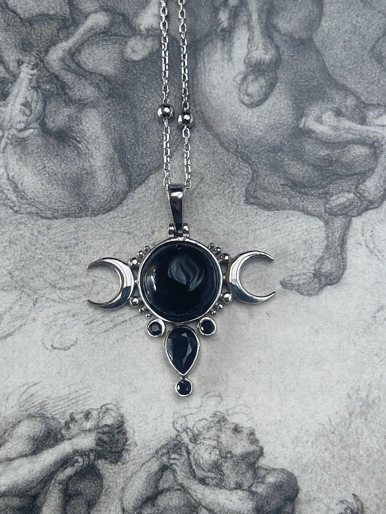 May include: Silver-toned pendant necklace featuring a central black gemstone, flanked by crescent moons. Additional black gemstones are set below the central stone. The necklace hangs on a delicate silver chain, creating a celestial-inspired design.