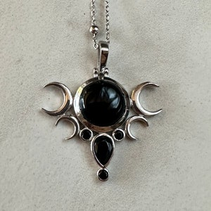 May include: Silver-toned pendant necklace featuring a central black gemstone surrounded by crescent moon and circular accents. A teardrop-shaped black stone hangs below. The necklace has a delicate chain with small, round beads.