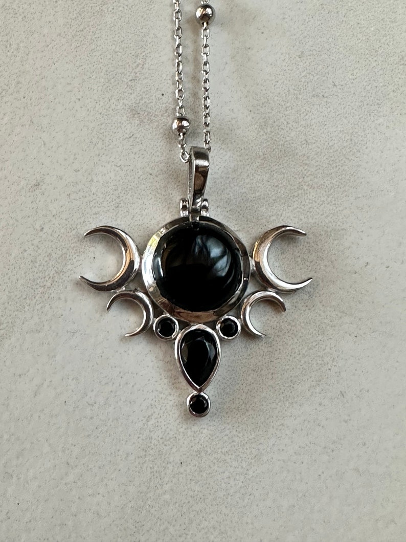May include: Silver-toned pendant necklace featuring a central black gemstone surrounded by crescent moon and circular accents. A teardrop-shaped black stone hangs below. The necklace has a delicate chain with small, round beads.