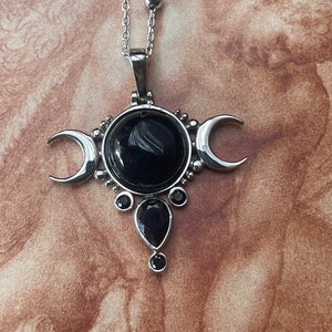 May include: Silver-toned pendant necklace featuring a central black gemstone, flanked by crescent moons. Additional black teardrop and round gemstones adorn the pendant. The necklace has a delicate chain with small, round beads.