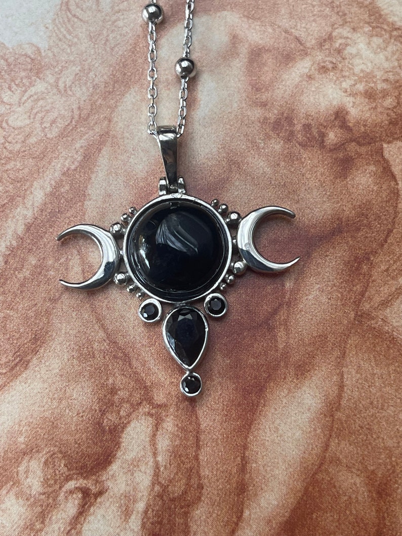May include: Silver-toned pendant necklace featuring a central black gemstone, flanked by crescent moons. Additional black teardrop and round gemstones adorn the pendant. The necklace has a delicate chain with small, round beads.