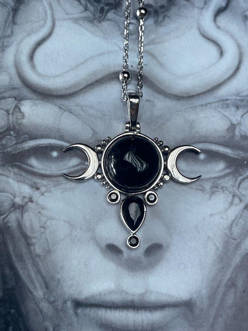 May include: Silver-toned pendant necklace featuring a central black stone surrounded by smaller black gems and crescent moon accents. The necklace has a delicate chain and a pendant with a gothic or celestial theme.