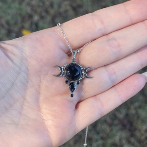 May include: Silver-toned necklace featuring a pendant with a large, round black stone at the center, flanked by crescent moon shapes. Additional black teardrop-shaped stones are below. The necklace is held in a hand against a blurred green background.