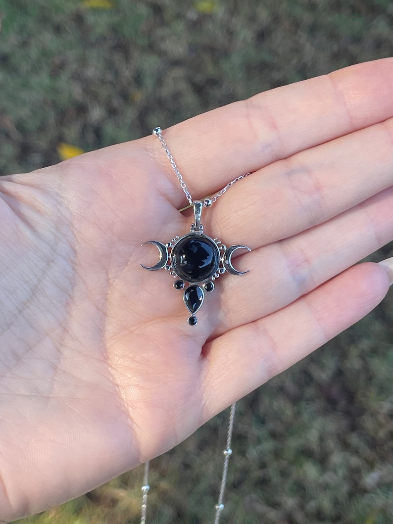 May include: Silver-toned necklace featuring a pendant with a large, round black stone at the center, flanked by crescent moon shapes. Additional black teardrop-shaped stones are below. The necklace is held in a hand against a blurred green background.
