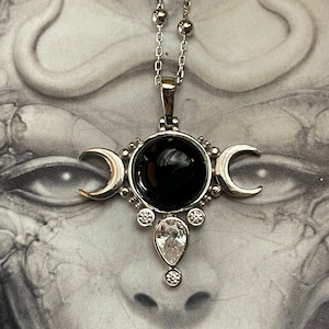 May include: Silver-toned pendant necklace featuring a central black stone surrounded by silver accents and crescent moon shapes. A teardrop-shaped clear stone hangs below the central stone. The necklace has a delicate chain with small silver beads.