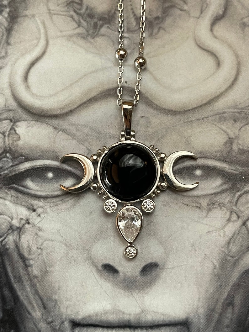 May include: Silver-toned pendant necklace featuring a central black stone surrounded by silver accents and crescent moon shapes. A teardrop-shaped clear stone hangs below the central stone. The necklace has a delicate chain with small silver beads.