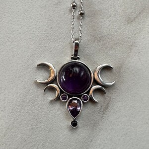 May include: A silver pendant necklace with a large, round, deep purple gemstone at the centre, framed by three crescent moons. Smaller purple gemstones and a teardrop-shaped stone are below. The chain is delicate and silver.