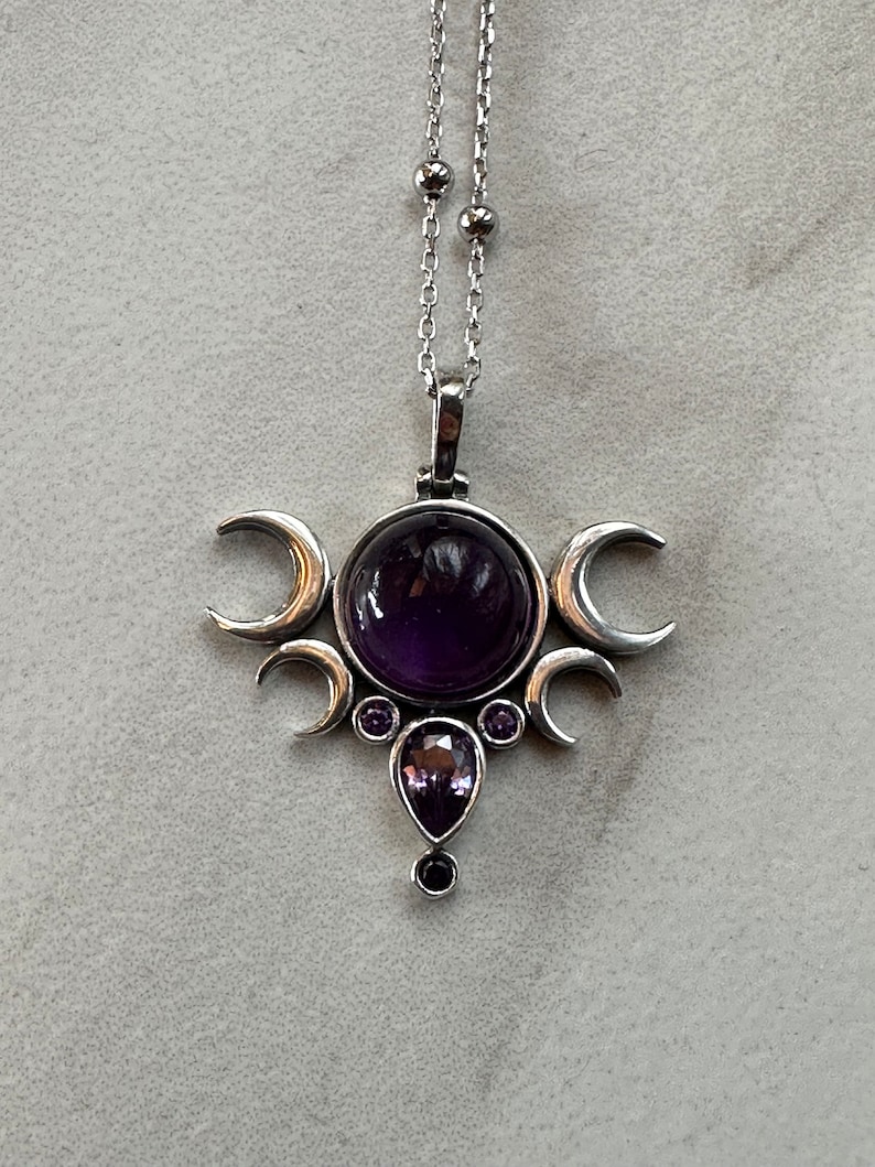 May include: A silver pendant necklace with a large, round, deep purple gemstone at the centre, framed by three crescent moons. Smaller purple gemstones and a teardrop-shaped stone are below. The chain is delicate and silver.