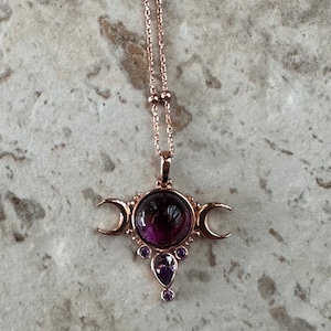 May include: A rose gold necklace with a pendant. The pendant has a large, round, dark purple gemstone at its centre, with crescent moons on either side. Smaller purple gemstones are below. The chain is delicate with small beads.