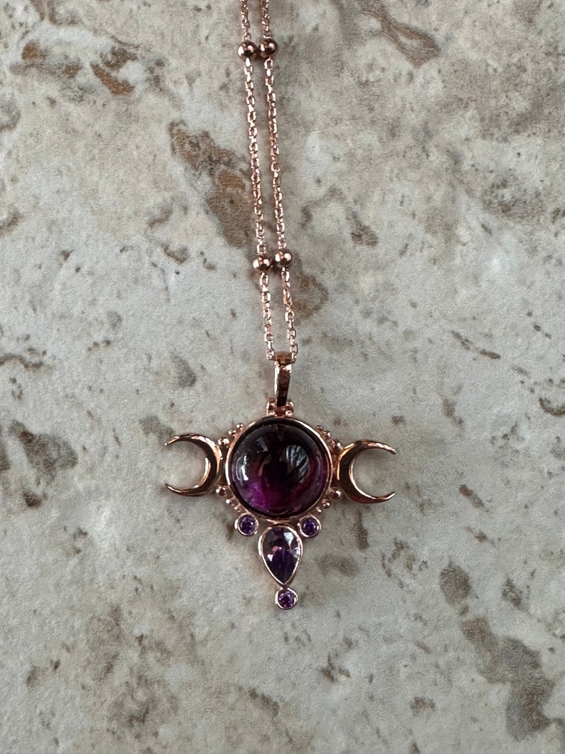 May include: A rose gold necklace with a pendant. The pendant has a large, round, dark purple gemstone at its centre, with crescent moons on either side. Smaller purple gemstones are below. The chain is delicate with small beads.