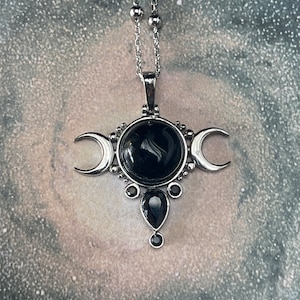 May include: Silver-toned pendant necklace featuring a central black gemstone surrounded by crescent moon accents. The pendant has a teardrop-shaped stone below the central stone, and is suspended from a delicate chain.