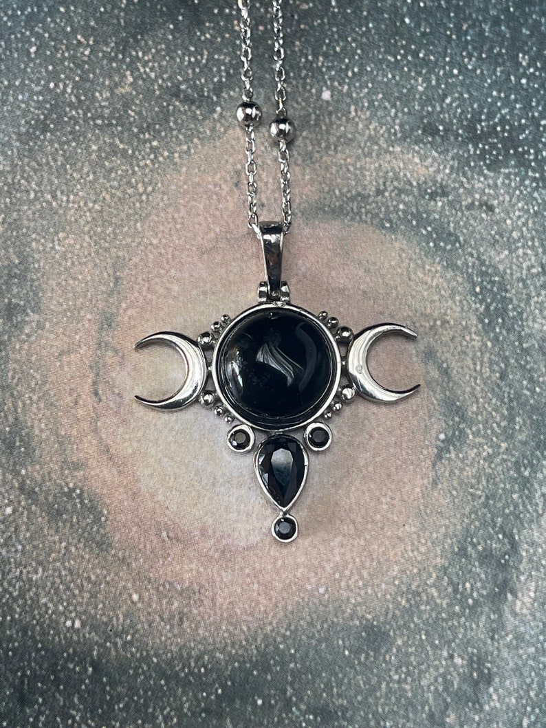 May include: Silver-toned pendant necklace featuring a central black gemstone surrounded by crescent moon accents. The pendant has a teardrop-shaped stone below the central stone, and is suspended from a delicate chain.