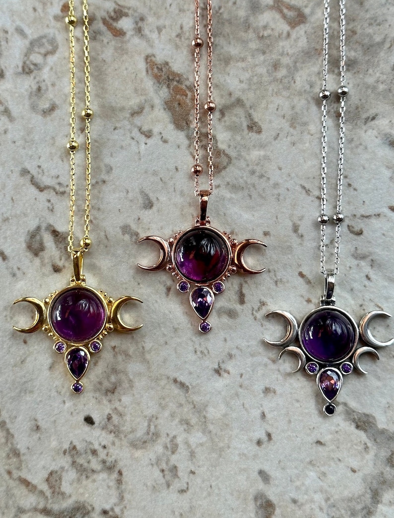 May include: Three necklaces with celestial pendants. The pendants feature a large, round, purple gemstone, crescent moon accents, and smaller purple and black gemstones. The chains are gold, rose gold, and silver.