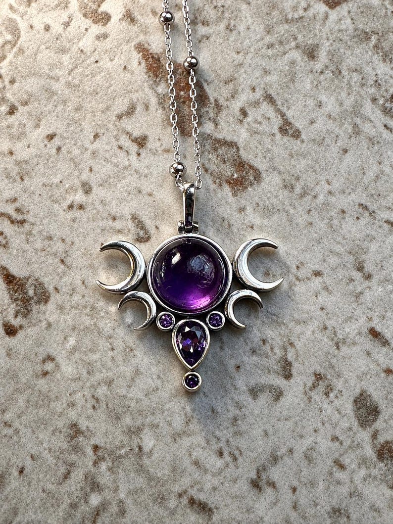 May include: A silver pendant necklace with a large, round, deep purple gemstone at its centre. The pendant features a triple moon design with crescent moons and smaller purple gemstones. The necklace chain is delicate.