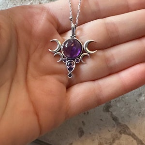 May include: A silver pendant necklace with a large, round, purple amethyst gemstone at the centre. The pendant has a crescent moon design and smaller purple gemstones. The necklace is displayed on a hand.