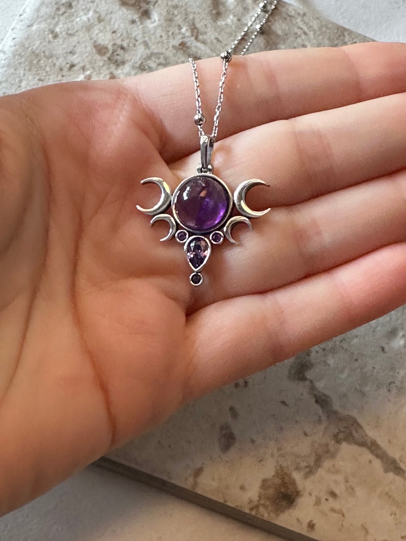 May include: A silver pendant necklace with a large, round, purple amethyst gemstone at the centre. The pendant has a crescent moon design and smaller purple gemstones. The necklace is displayed on a hand.