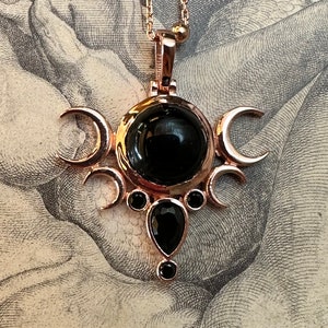 May include: Rose gold-toned pendant necklace featuring a central black gemstone surrounded by crescent moon shapes and smaller black gems. The pendant hangs from a delicate chain. The design has a celestial theme.
