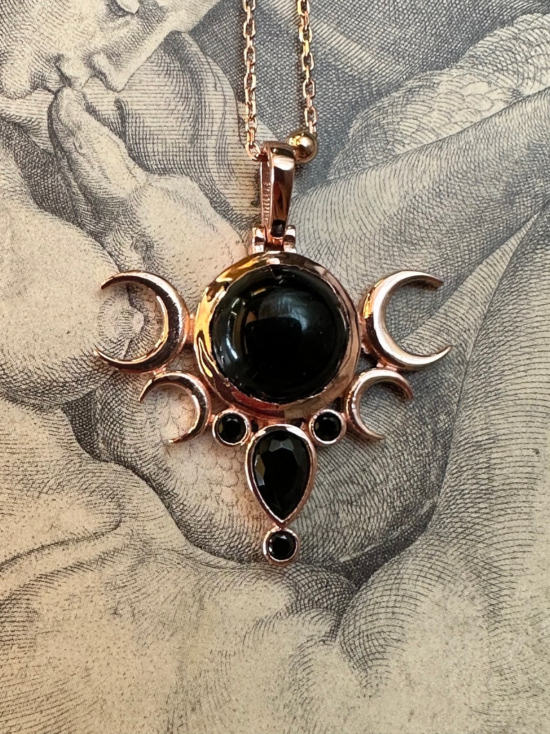 May include: Rose gold-toned pendant necklace featuring a central black gemstone surrounded by crescent moon shapes and smaller black gems. The pendant hangs from a delicate chain. The design has a celestial theme.