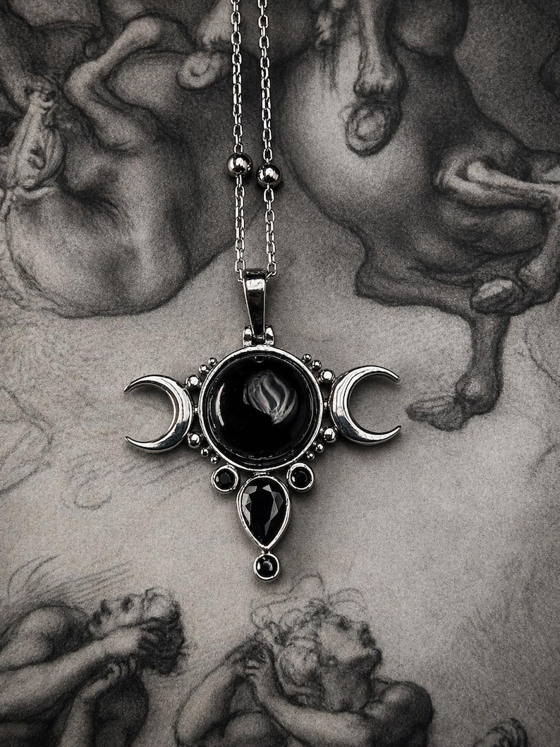 May include: A silver-toned pendant necklace featuring a central black gemstone surrounded by crescent moon accents. The pendant has a teardrop-shaped black gem at the bottom and hangs from a delicate chain. The background is a grayscale drawing.
