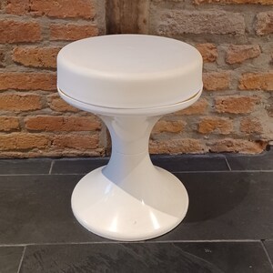 Könnte beinhalten: Ein weißer, runder Hocker aus Kunststoff mit einer breiten Basis. Der Hocker steht auf einem schwarzen Fliesenboden.