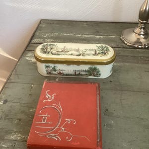 Könnte beinhalten: Zwei Vintage-Boxen, eine mit weißem Hintergrund und goldener Verzierung mit einer gemalten Szene einer Stadt, und die andere eine rote Box mit silbernem Design.