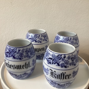 Könnte beinhalten: Vier weiße Keramikkanister mit blau-weißen Blumendesigns. Jeder Kanister trägt eine schwarze Aufschrift: "Griesmehl", "Kaffee", "Suppen" und "Zucker".