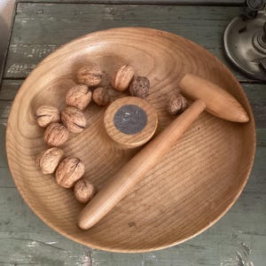Könnte beinhalten: Eine Holzschale gefüllt mit Walnüssen, einem Holz-Nussknacker und einem Werkzeug zum Knacken von Nüssen. Die Schale und die Werkzeuge sind aus hellem Holz gefertigt. Der Nussknacker hat einen abgerundeten Griff und einen keilförmigen Kopf. Das Nussknackwerkzeug ist rund mit dunkler Mitte.