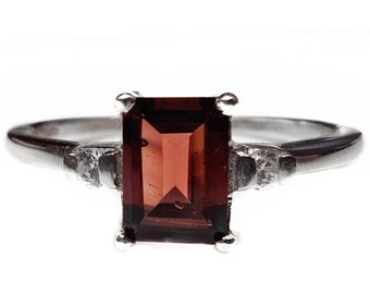 May include: A silver ring with a dark red rectangular gemstone and two smaller clear gemstones on the band.