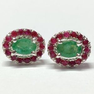 Może przedstawiać: Para srebrnych kolczyków z owalnymi zielonymi kamieniami szlachetnymi otoczonymi halo z czerwonych kamieni szlachetnych.