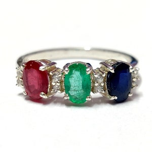 Peut inclure: Une bague en argent avec trois pierres précieuses de forme ovale : un rubis rouge, une émeraude verte et un saphir bleu. La bague est sertie de petits diamants autour des pierres précieuses.