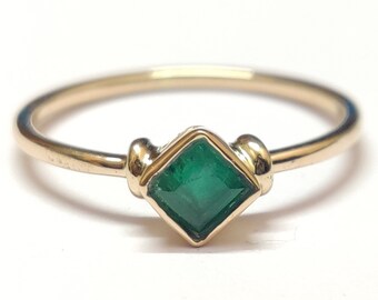 May include: A gold ring with a square emerald gemstone set in the center. The ring has a simple band and the emerald is a vibrant green color.