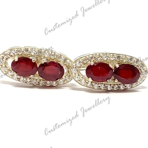 May include: A pair of oval-shaped earrings with a gold setting and two red gemstones surrounded by a halo of smaller white gemstones.