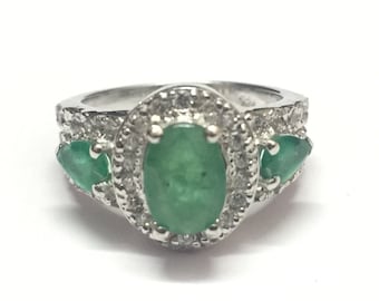 May include: A silver ring with a large oval emerald gemstone surrounded by smaller diamonds. Two smaller emerald gemstones are on either side of the center stone.
