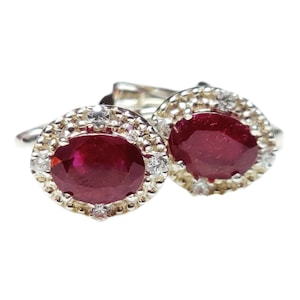 Op de afbeelding: Een paar zilveren manchetknopen met ovale, dieprode edelstenen, omringd door een halo van kleinere, heldere stenen. Klassiek ontwerp, geschikt voor formele gelegenheden.