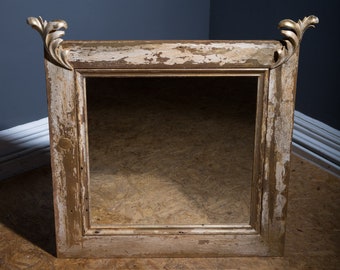 May include: A square mirror with a distressed gold-painted wooden frame. The frame features ornate carved leaf-shaped finials at the top corners and shows significant paint wear, revealing the underlying wood texture throughout.