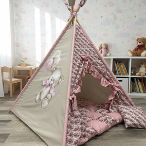 Könnte beinhalten: Ein beiges und rosa Tipi-Zelt für Kinder. Die Vorderseite zeigt eine Illustration von Teddybär und Hase. Der Innenraum ist mit einem rosa gemusterten Stoff ausgekleidet, und der Eingang ist mit Rüschen verziert. Das Zelt steht in einem Kinderzimmer.
