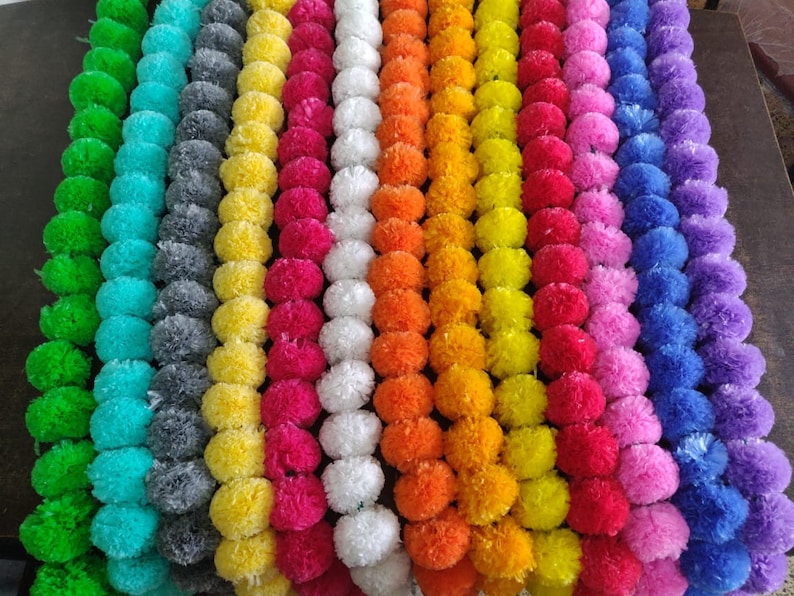 K&ouml;nnte beinhalten: Eine bunte Girlande aus Marigolden-Bl&uuml;tenpompons. Die Girlande besteht aus mehreren Str&auml;ngen von Pompons in verschiedenen Farben, darunter Gr&uuml;n, T&uuml;rkis, Grau, Gelb, Rosa, Orange, Rot und Lila.