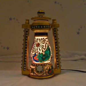 Op de afbeelding: Een witte marmeren lantaarn met ingewikkelde gouden en kleurrijke beschilderde details, met een pauwenmotief. De lantaarn heeft een lichtje erin, waardoor het ontwerp wordt verlicht.