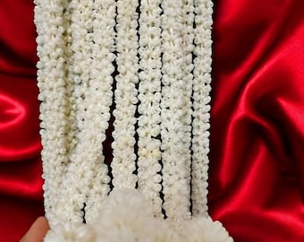 Op de afbeelding: Een witte krans gemaakt van kleine, delicate bloemen. De krans is in meerdere strengen gerangschikt en is gedrapeerd over een rood oppervlak.