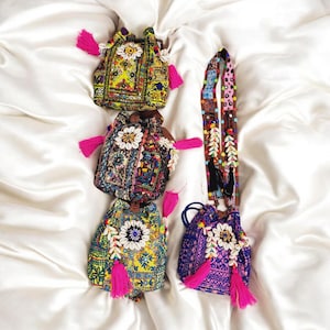 Puede incluir: Cuatro bolsas de cordón de colores con borlas rosas. Cada bolsa está decorada con bordados y cuentas. Las bolsas están dispuestas sobre un fondo de tela blanca.